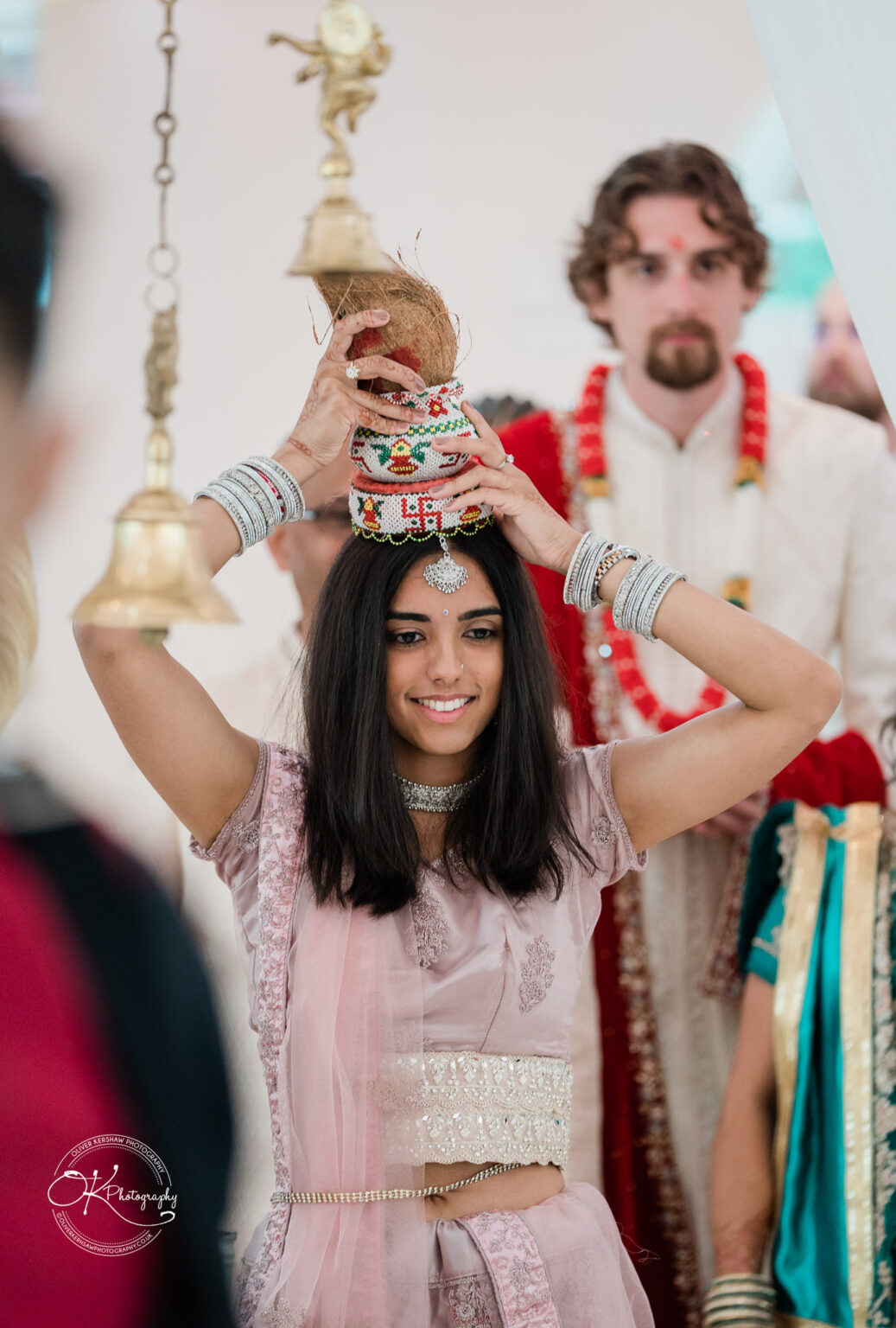 A bride in a traditional outfit holds a decorative pot above her head during a wedding ceremony at Winstanley House, with guests in the background.