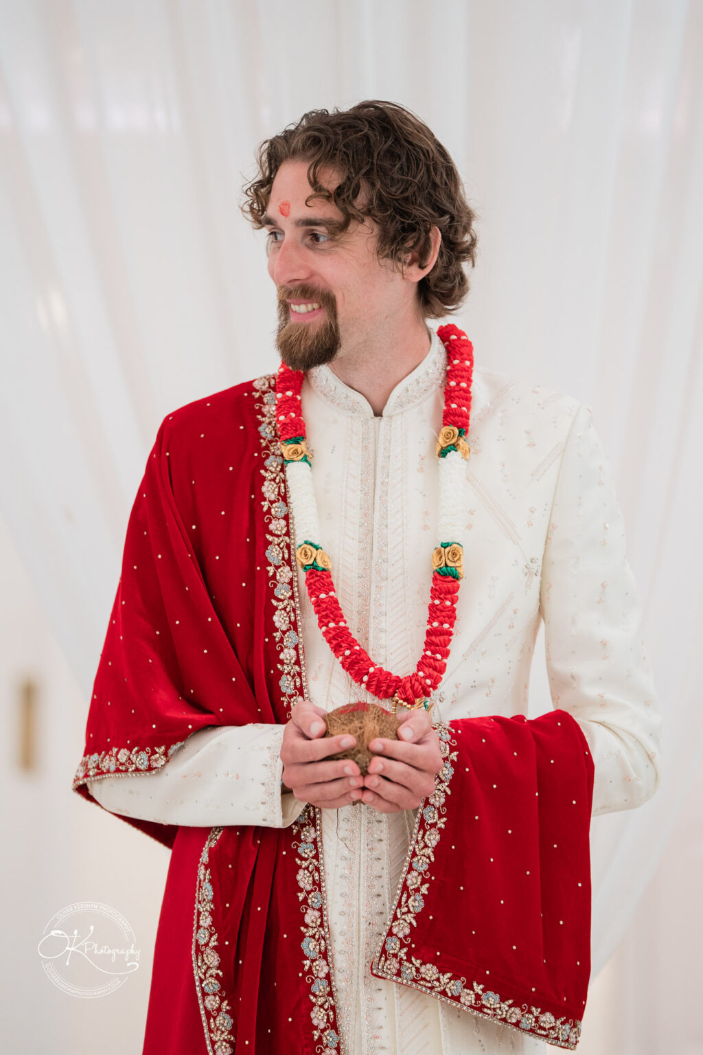 A groom in a traditional white and red outfit stands holding a ceremonial object, adorned with a floral garland and a decorative shawl.