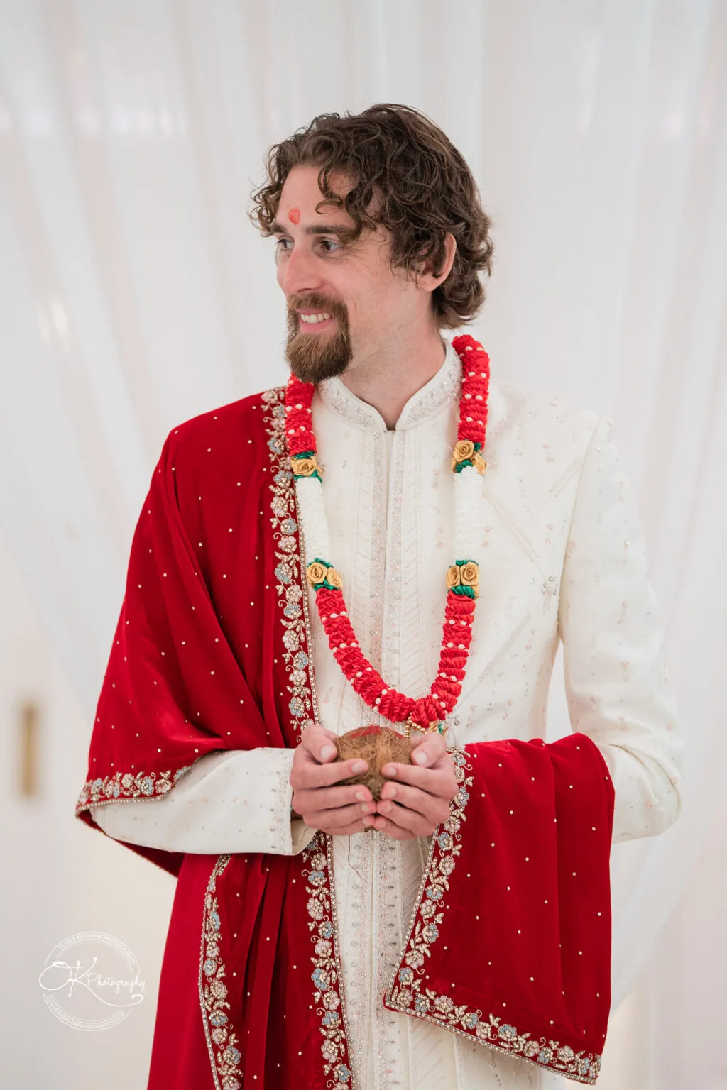 Roshni & Joseph-wedding -Winstanley House-Photography By Oliver Kershaw - OK904189 A groom in a traditional white and red outfit stands holding a ceremonial object, adorned with a floral garland and a decorative shawl.