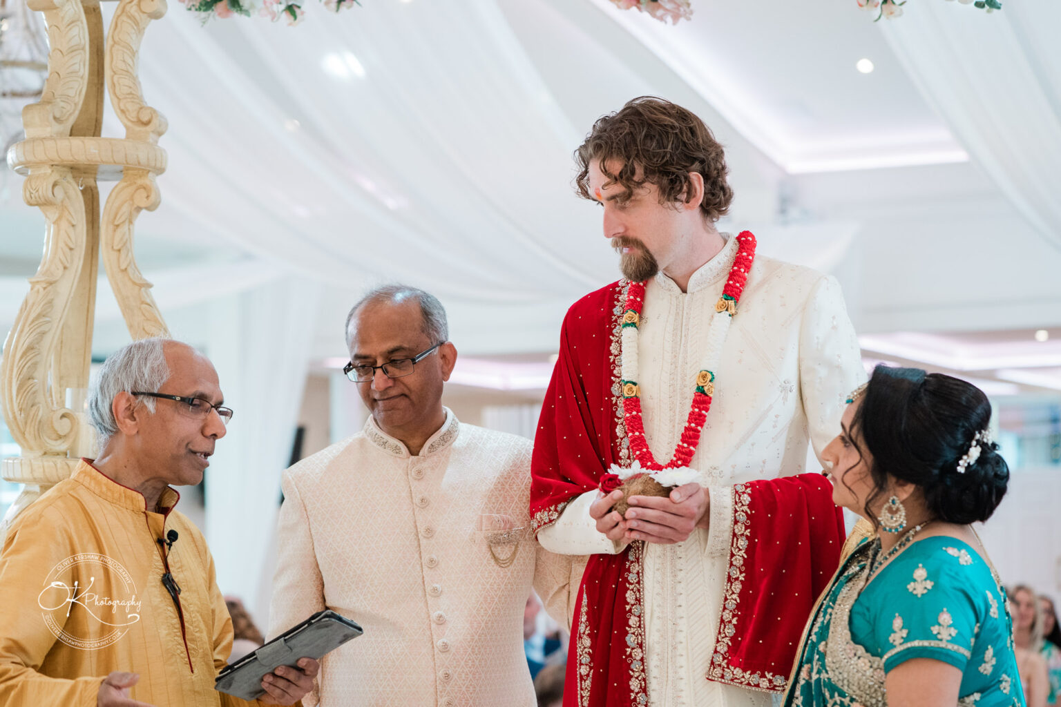 A groom in traditional attire stands with a bride in a turquoise outfit, while two officiants address them under an ornate arch.