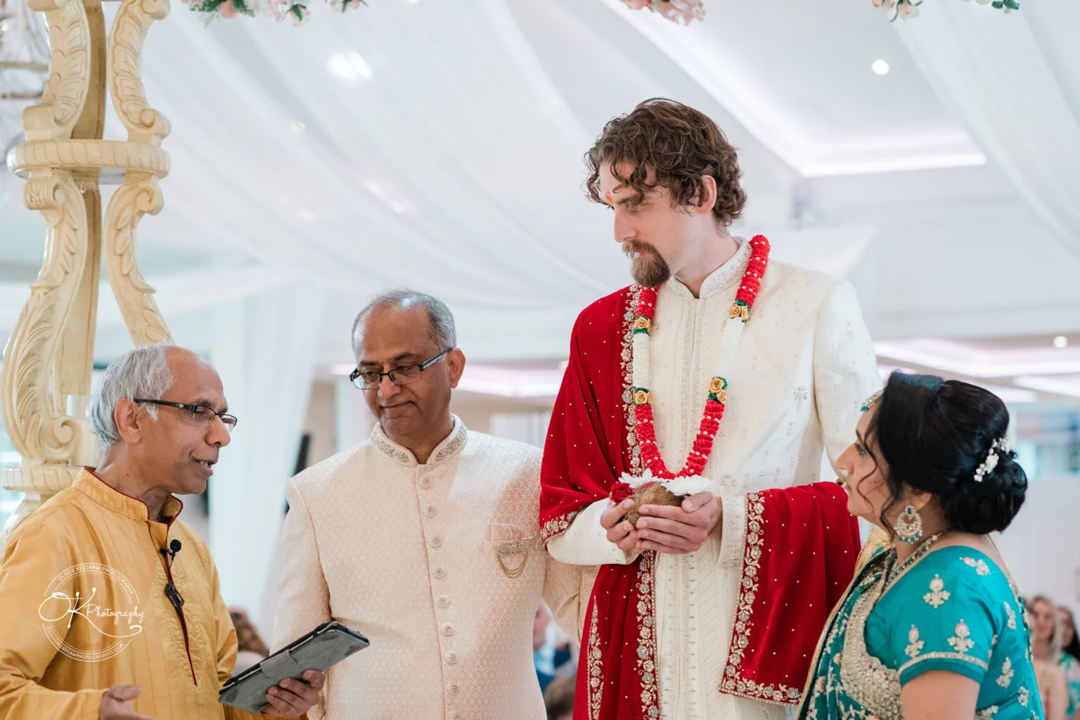 Roshni & Joseph-wedding -Winstanley House-Photography By Oliver Kershaw - OK904299 A groom in traditional attire stands with a bride in a turquoise outfit, while two officiants address them under an ornate arch.