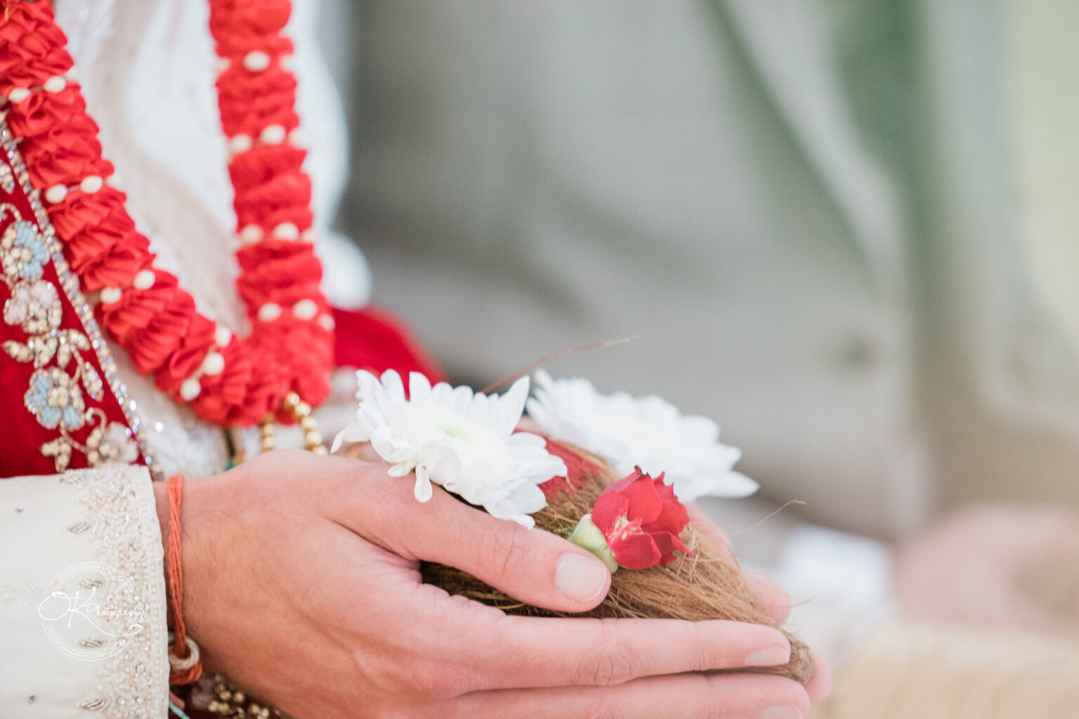 A close-up of a hand holding a small bouquet of white and red flowers, adorned with decorative clothing in a wedding setting.
