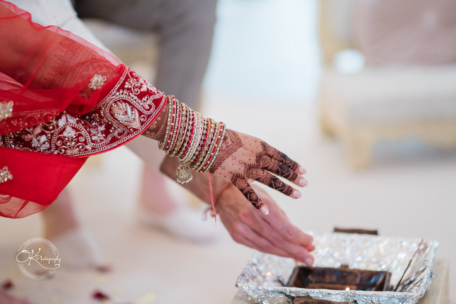 A close-up of a decorated hand adorned with intricate henna and bangles, reaching towards a ceremonial fire in a wedding setting.