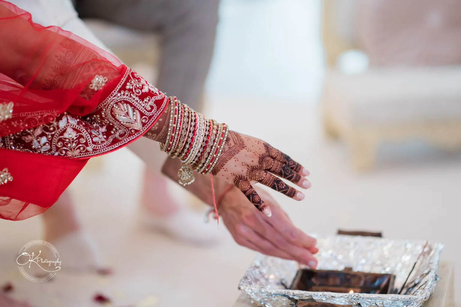 Roshni & Joseph-wedding -Winstanley House-Photography By Oliver Kershaw - OK904460 A close-up of a decorated hand adorned with intricate henna and bangles, reaching towards a ceremonial fire in a wedding setting.