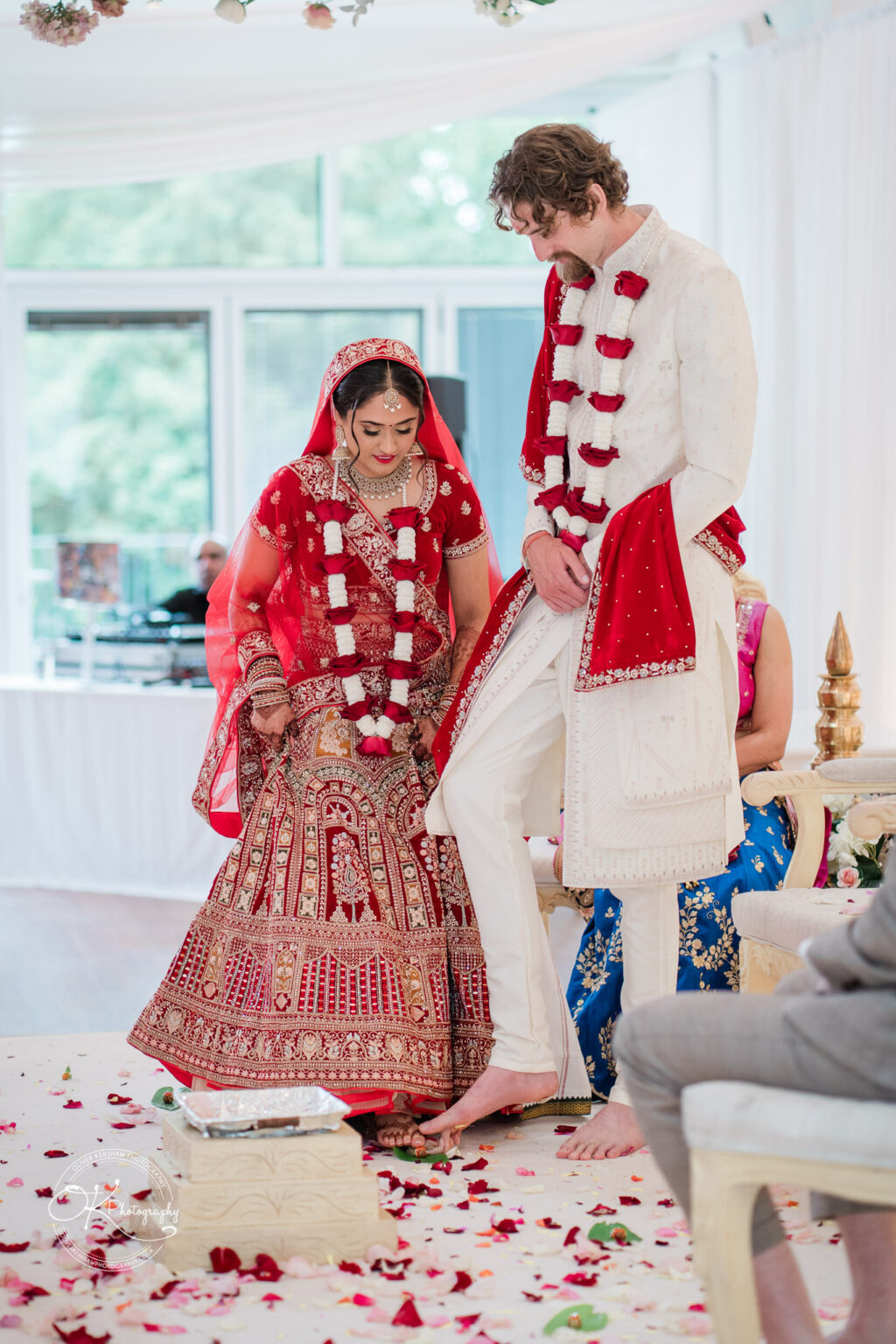A bride in a vibrant red and gold wedding dress with a matching dupatta, stands beside a tall groom in a white sherwani, during a wedding ceremony at Winstanley House. They are participating in a traditional ritual, with flower petals scattered around them.