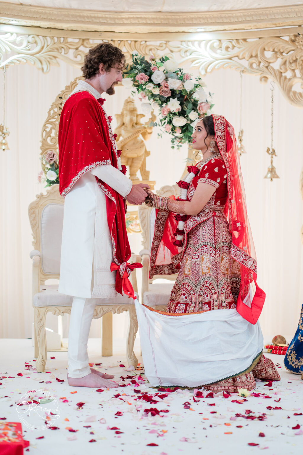 A bride in a red and gold traditional outfit holds hands with a groom in a white and red ensemble during a wedding ceremony at Winstanley House, surrounded by floral decorations.