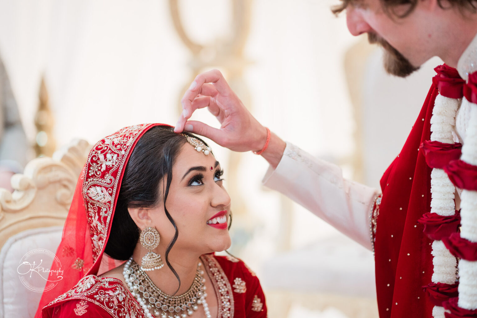 A bride in a red and gold wedding outfit smiles as her partner adjusts her headpiece during a ceremony at Winstanley House.