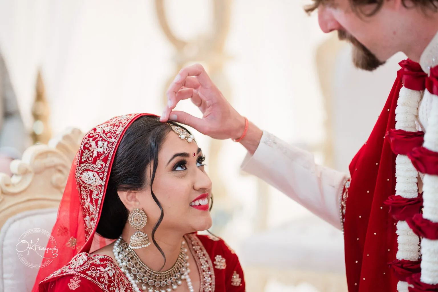 Roshni & Joseph-wedding -Winstanley House-Photography By Oliver Kershaw - OK904545 A bride in a red and gold wedding outfit smiles as her partner adjusts her headpiece during a ceremony at Winstanley House.