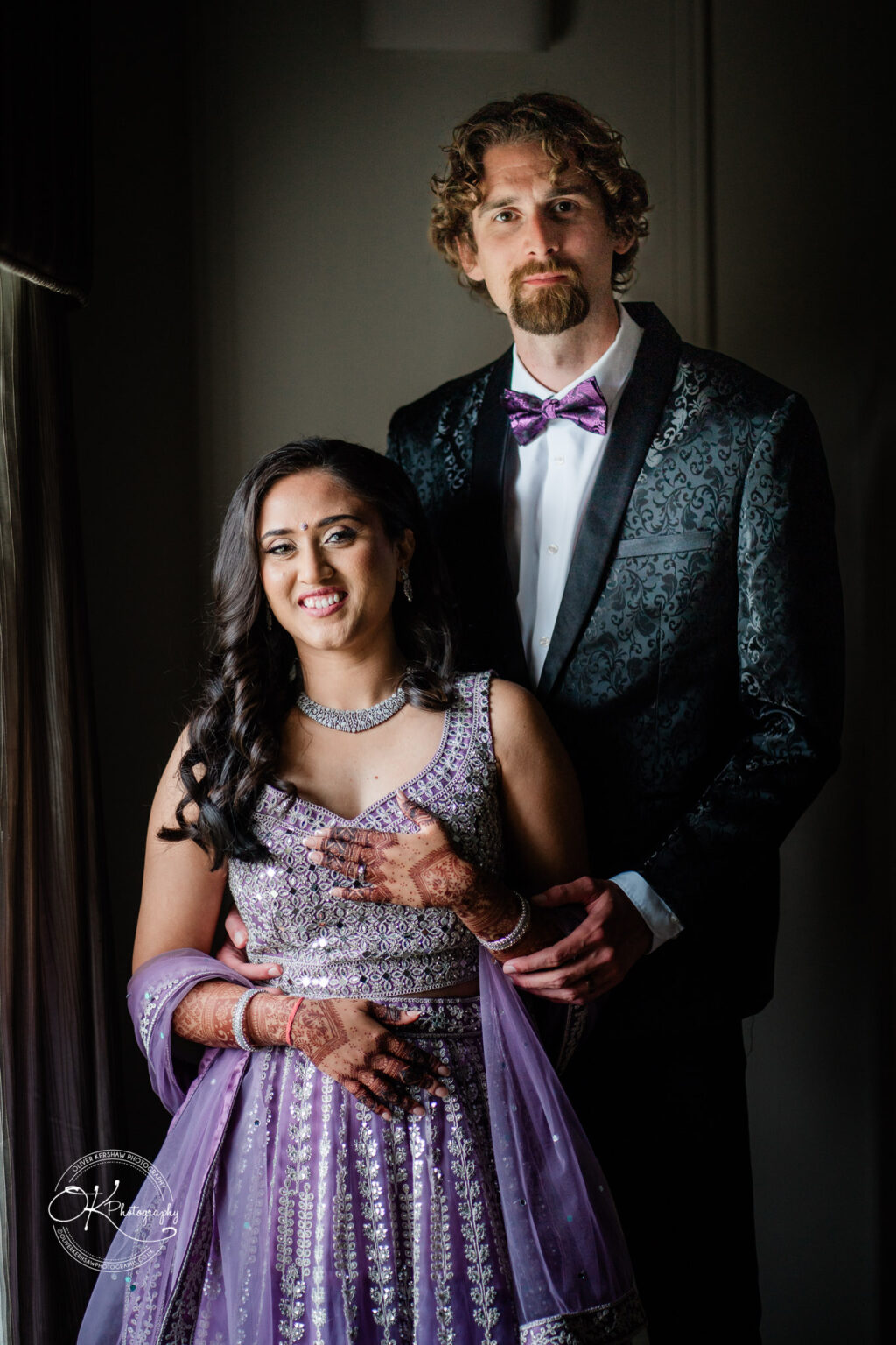A couple standing together, traditionally dressed; the woman in a purple lehenga with intricate patterns, and the man in a formal black suit with a bow tie.
