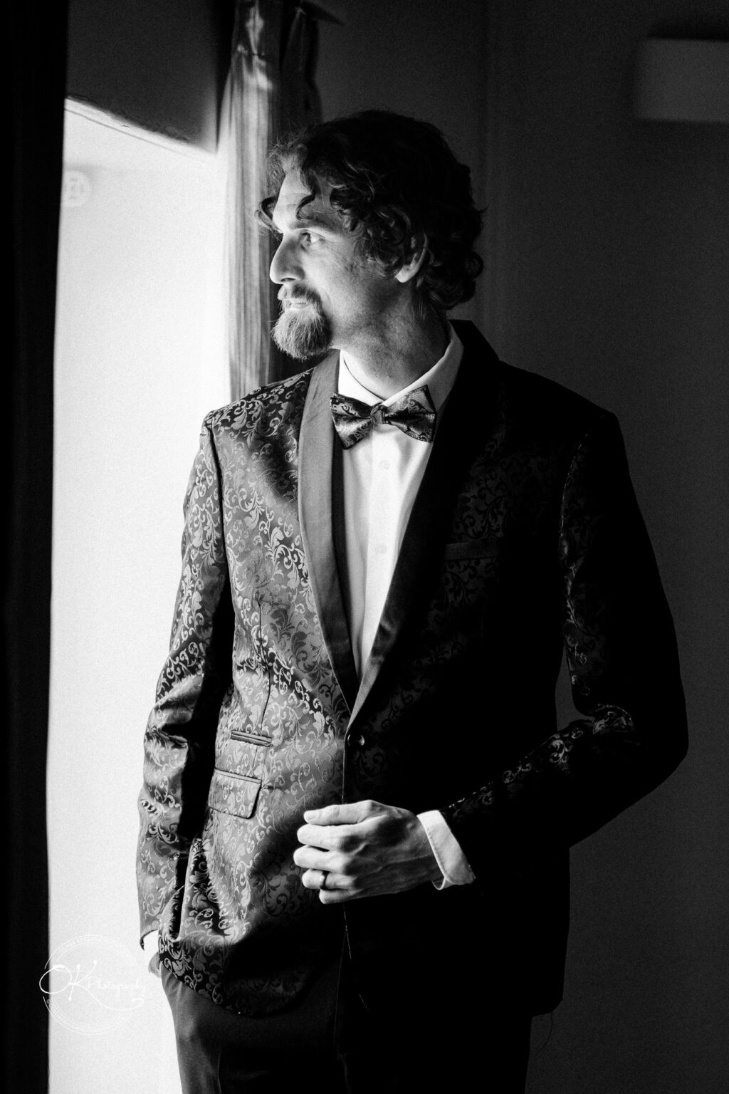 A man in a patterned black tuxedo and bow tie stands thoughtfully by a window, showcasing a stylish wedding look.