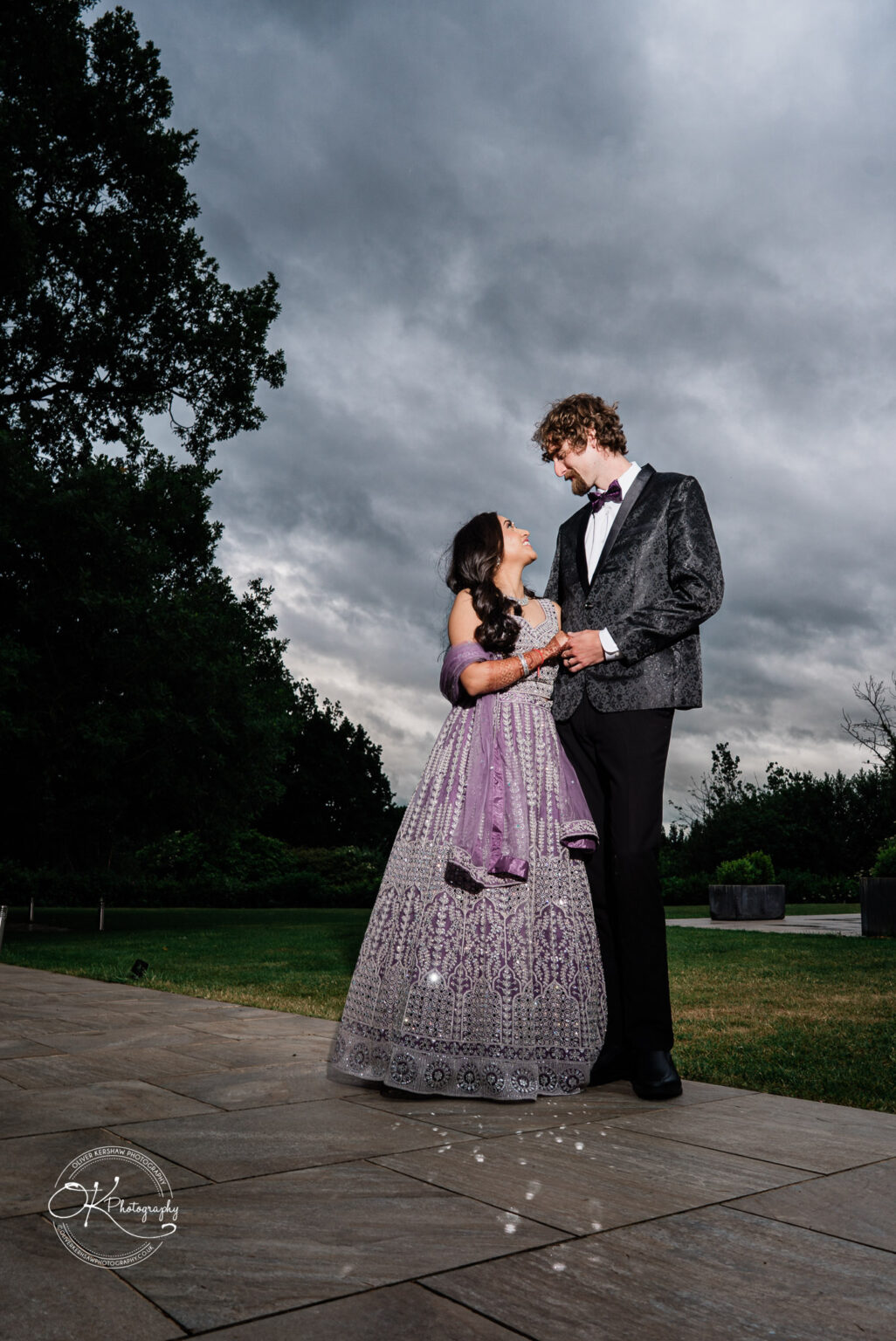 A couple dressed elegantly for a wedding, standing together outdoors, smiling at each other with dark, cloudy skies in the background.