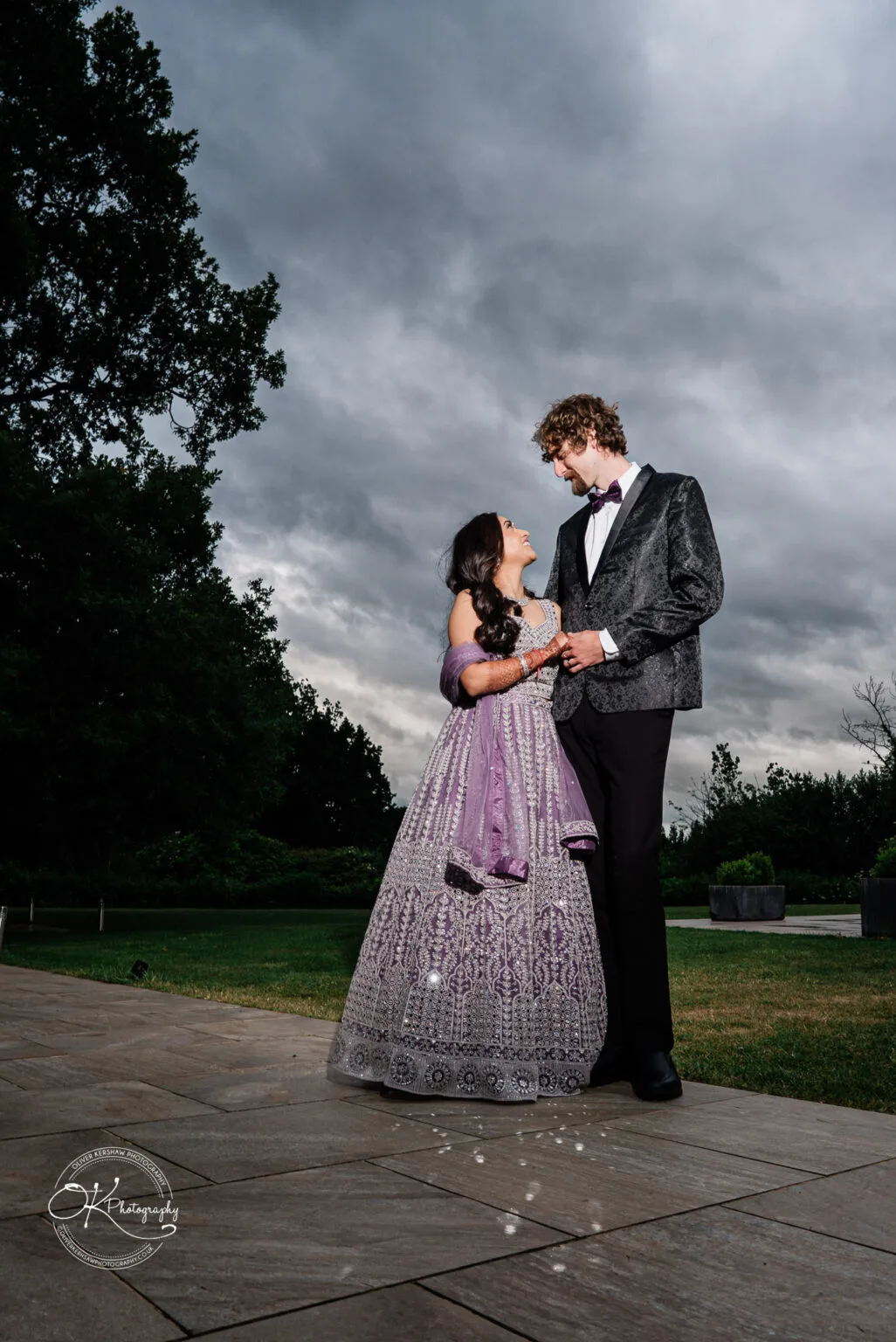 Roshni & Joseph-wedding -Winstanley House-Photography By Oliver Kershaw - OK904769 A couple dressed elegantly for a wedding, standing together outdoors, smiling at each other with dark, cloudy skies in the background.
