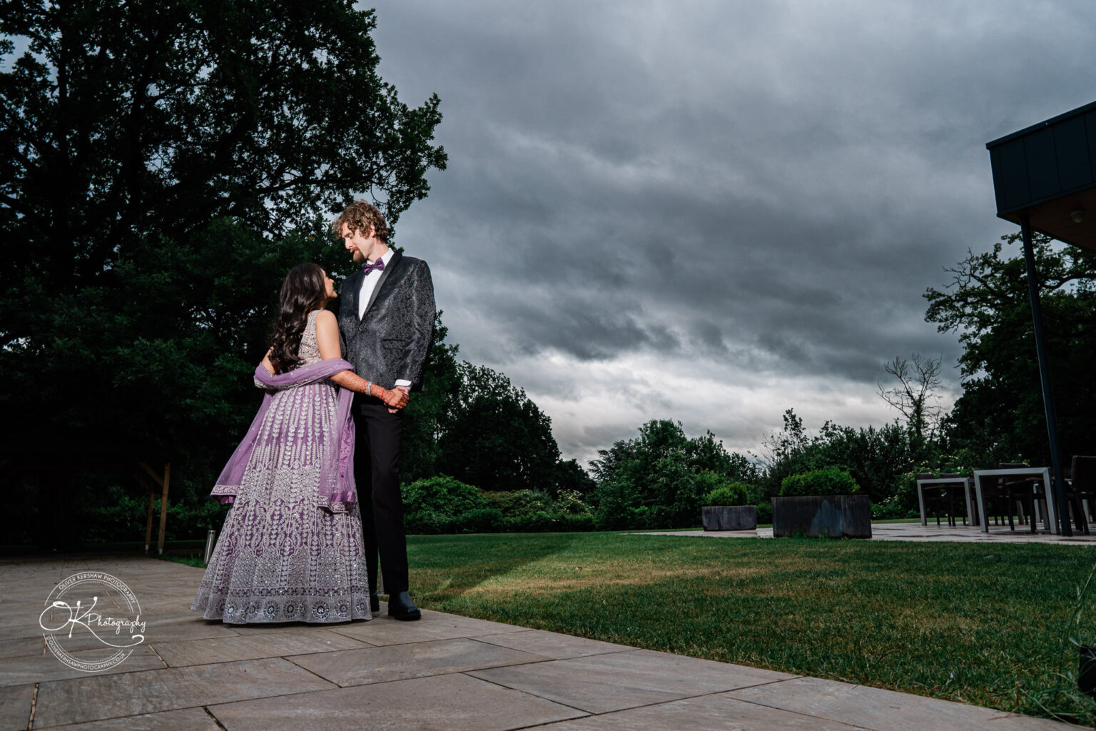 A couple stands close together in an outdoor setting, dressed elegantly, with dramatic clouds overhead.