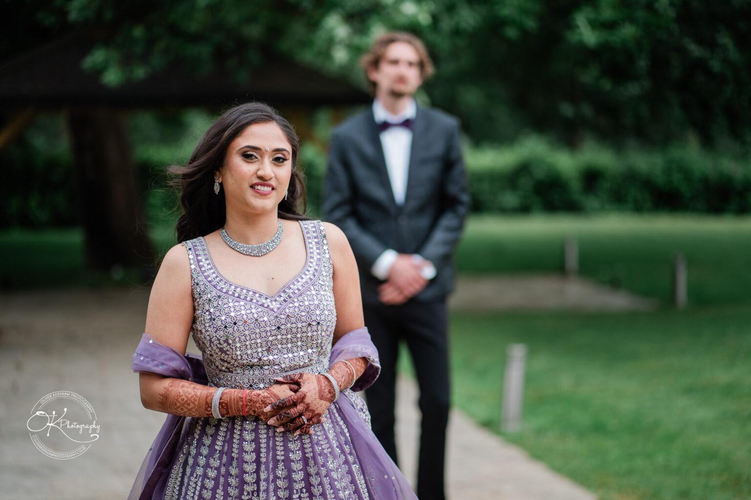 A bride in a purple embellished gown walks towards a groom in a suit against a lush garden backdrop at Winstanley House.