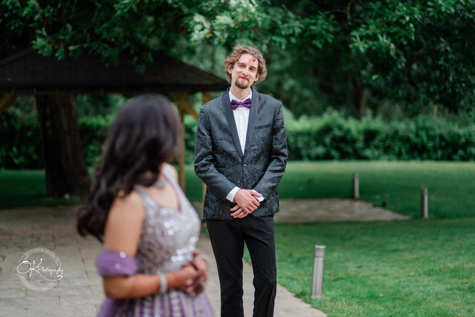 A groom in a black suit and bow tie stands smiling at a bride in a sparkly purple gown, with a green garden backdrop.