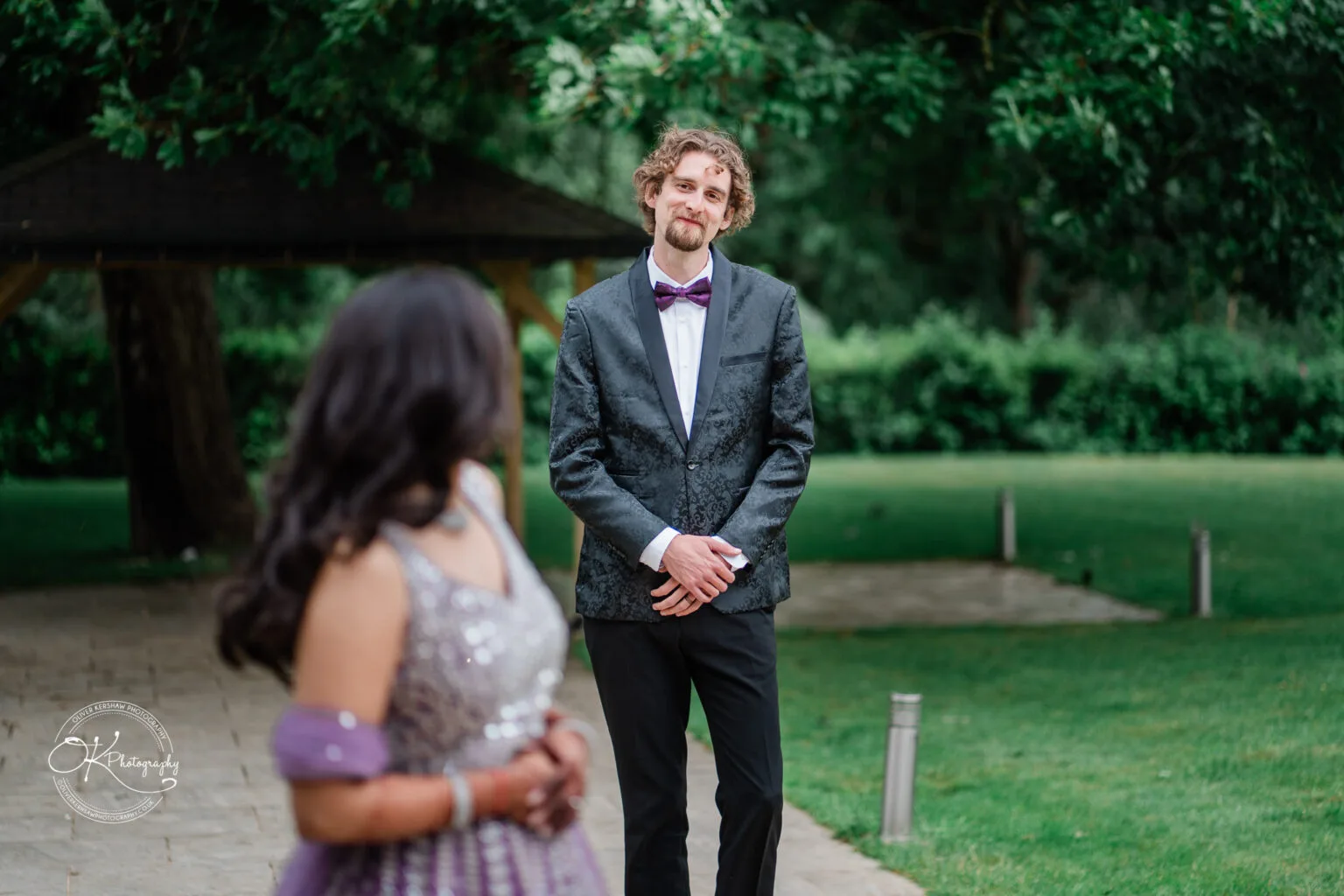 Roshni & Joseph-wedding -Winstanley House-Photography By Oliver Kershaw - OK904792 A groom in a black suit and bow tie stands smiling at a bride in a sparkly purple gown, with a green garden backdrop.