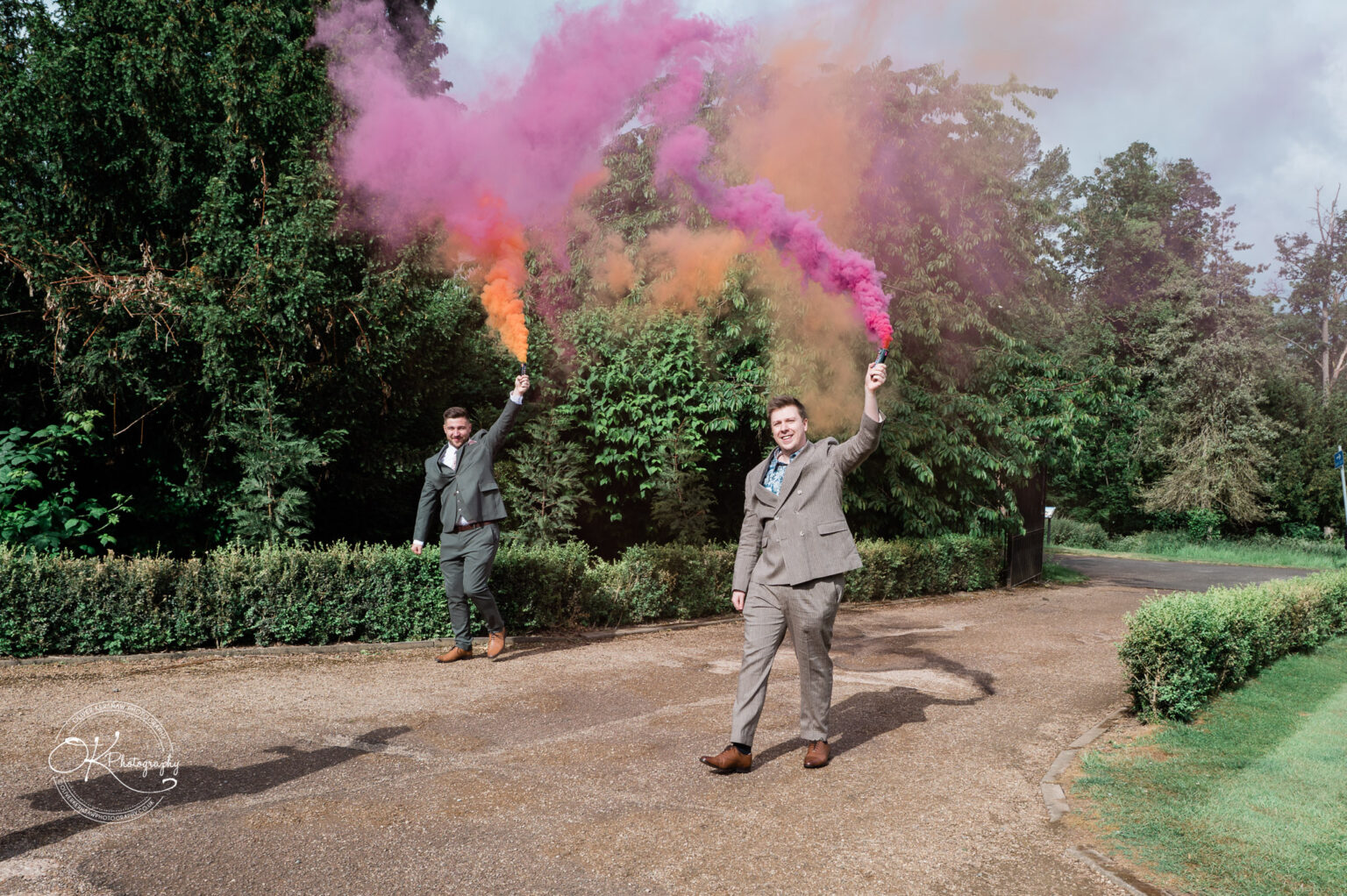 Two well-dressed men holding colorful smoke flares, walking along a path surrounded by greenery at Winstanley House.