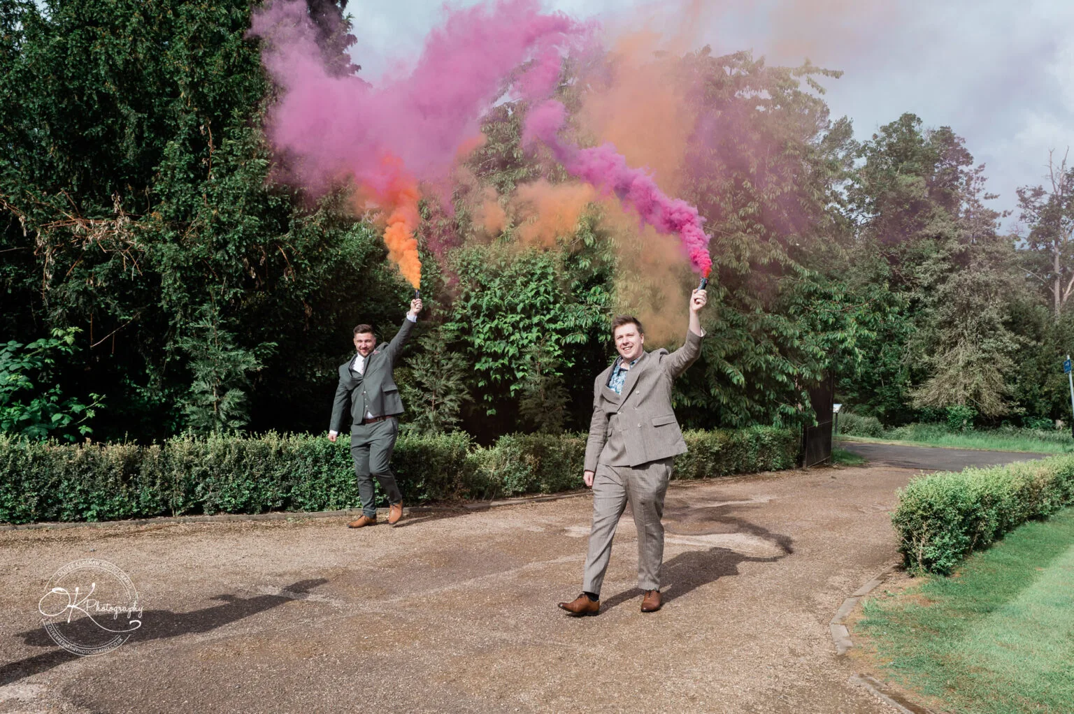Roshni & Joseph-wedding -Winstanley House-Photography By Oliver Kershaw - Z62_4983 Two well-dressed men holding colorful smoke flares, walking along a path surrounded by greenery at Winstanley House.
