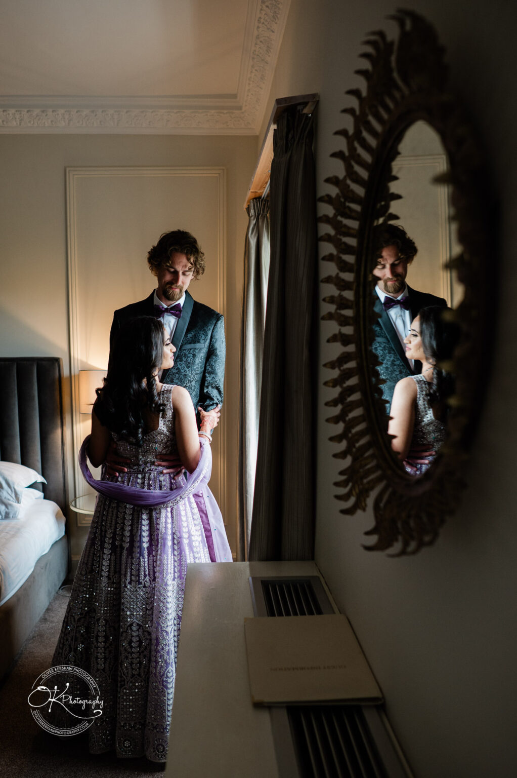 A bride and groom in elegant attire share a moment, reflected in a mirror, in a beautifully decorated room at Winstanley House.