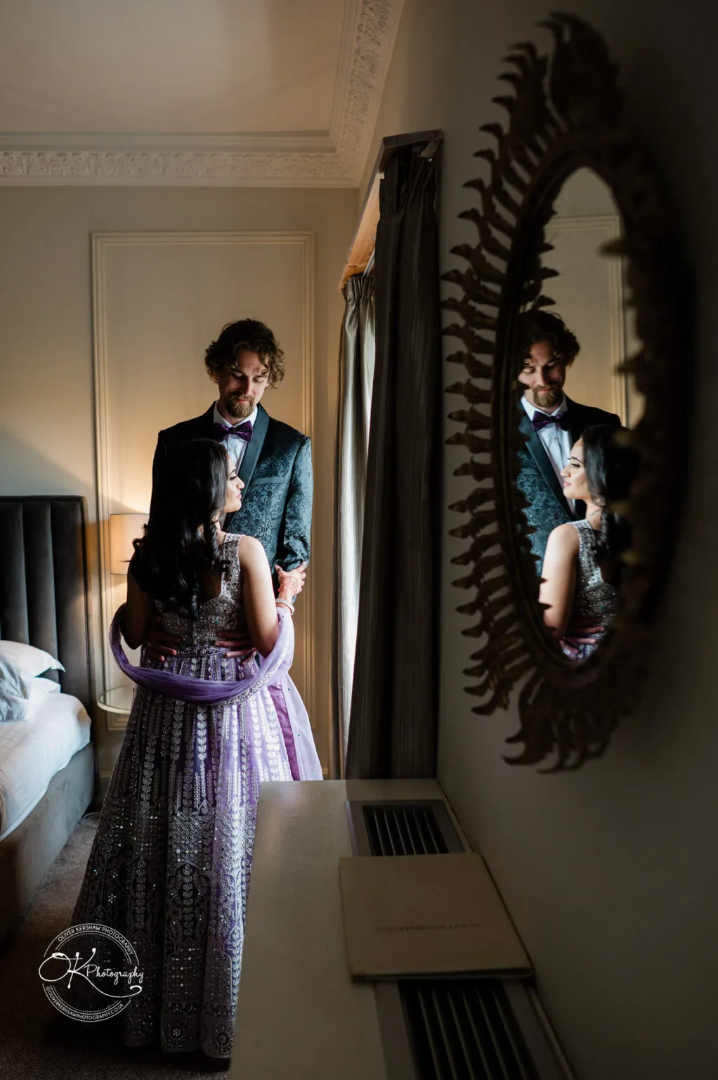 Roshni & Joseph-wedding -Winstanley House-Photography By Oliver Kershaw - Z62_5658 A bride and groom in elegant attire share a moment, reflected in a mirror, in a beautifully decorated room at Winstanley House.