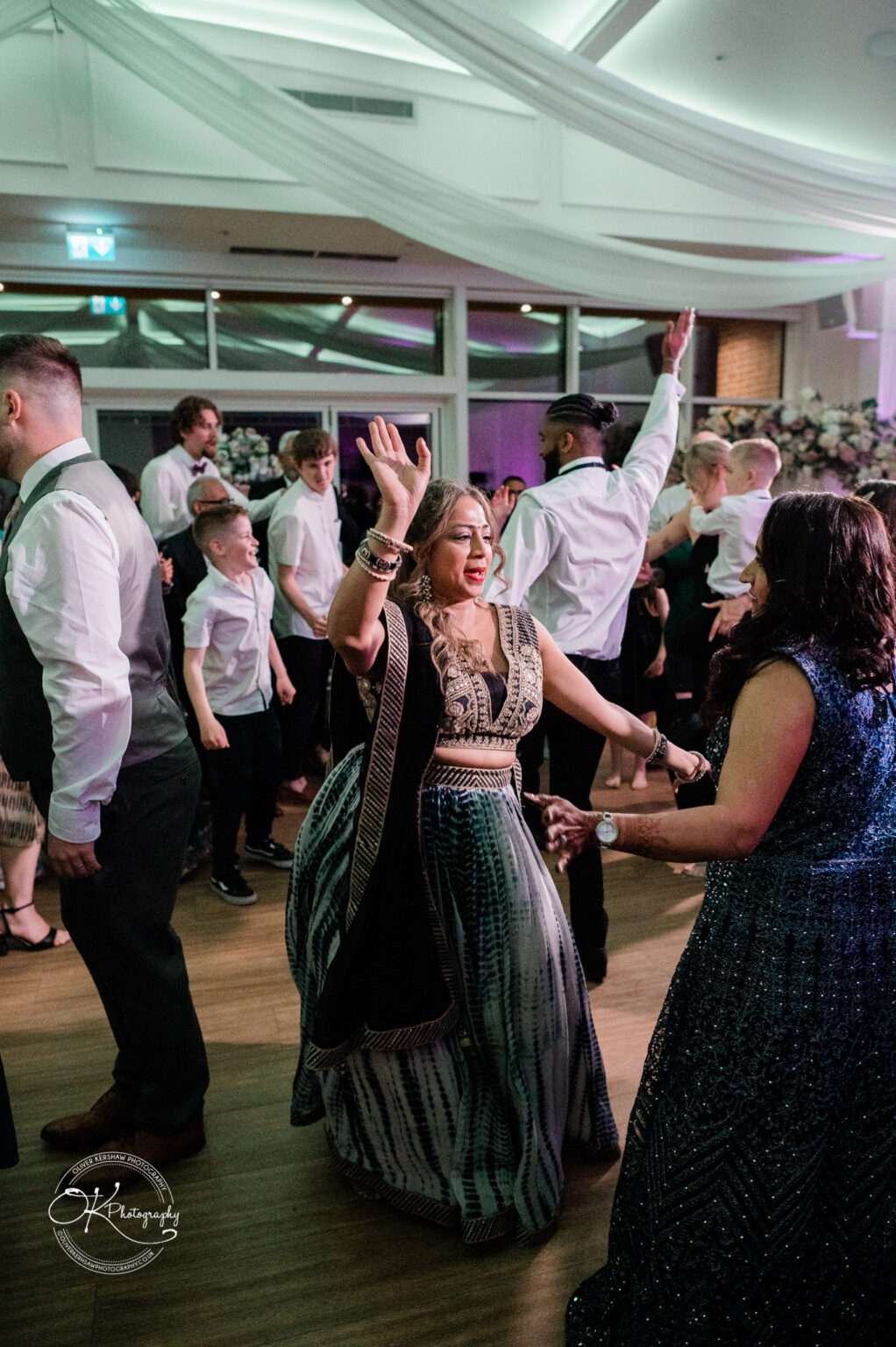 A joyful woman in a traditional dress dances with her hands raised, surrounded by guests at a wedding reception in a lively atmosphere.