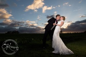 Bride and groom kissing outdoors at sunset with dramatic sky.