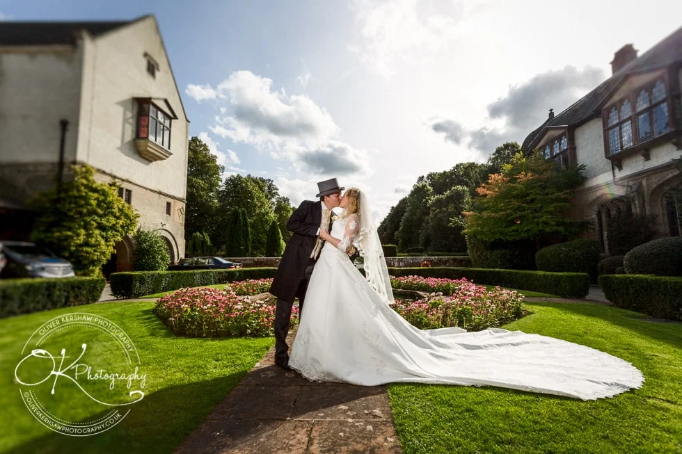 Bride and groom embrace and kiss in a garden with historic buildings and colourful flower beds in the background.