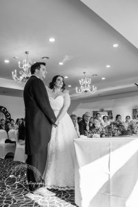 Bride and groom standing together during a wedding ceremony, with seated guests watching attentively in the background, in a chandelier-lit room.