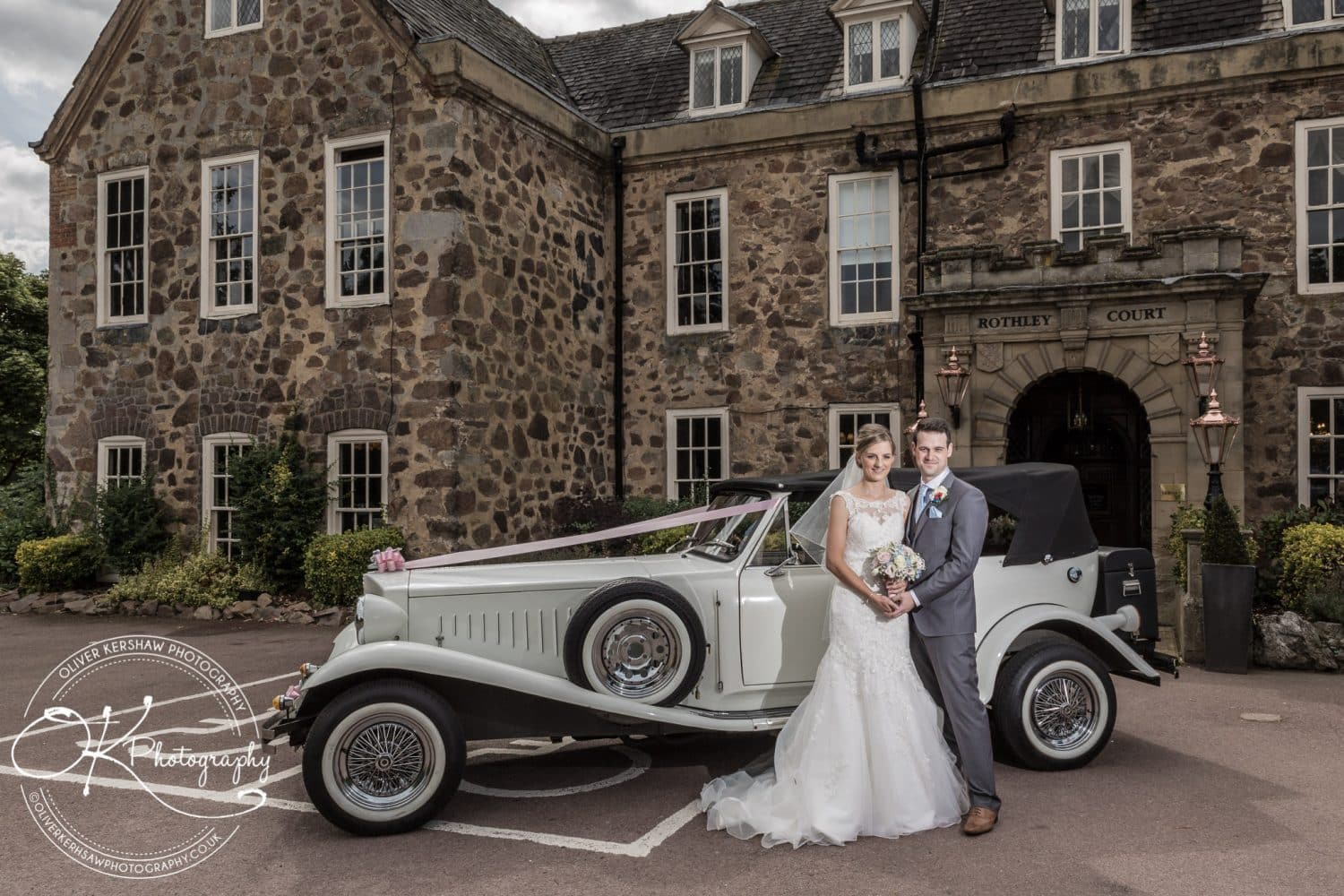 Bride and groom posing in front of a vintage car and historic stone building.